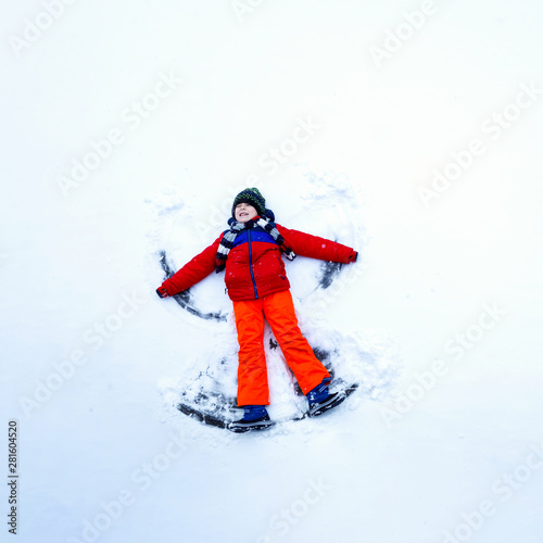 Cute little kid boy in colorful winter clothes making snow angel, laying down on snow. Active outdoors leisure with children in winter. Happy child having fun and laughing.