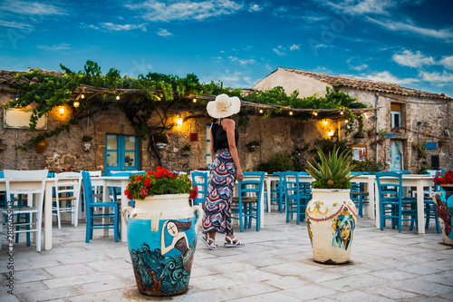 Young romantic woman walks near the colorful outdoor cafe  in the beautiful sicilian coastal village Marzamemi in Sicily, south Italy photo