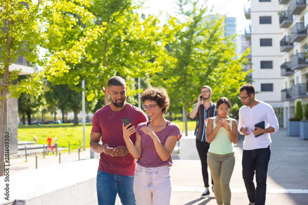 Obraz premium Happy young people using mobile phones while walking through residential area. Men and women talking on cell, using smartphones and showing screen to each other. Wi-Fi outdoors concept