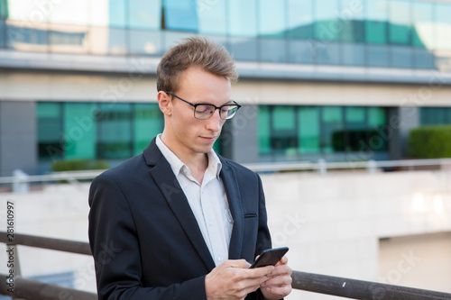Serious focused manager using online mobile app outdoors. Young business man in eyeglasses standing in urban settings and texting message on smartphone. Business communication concept