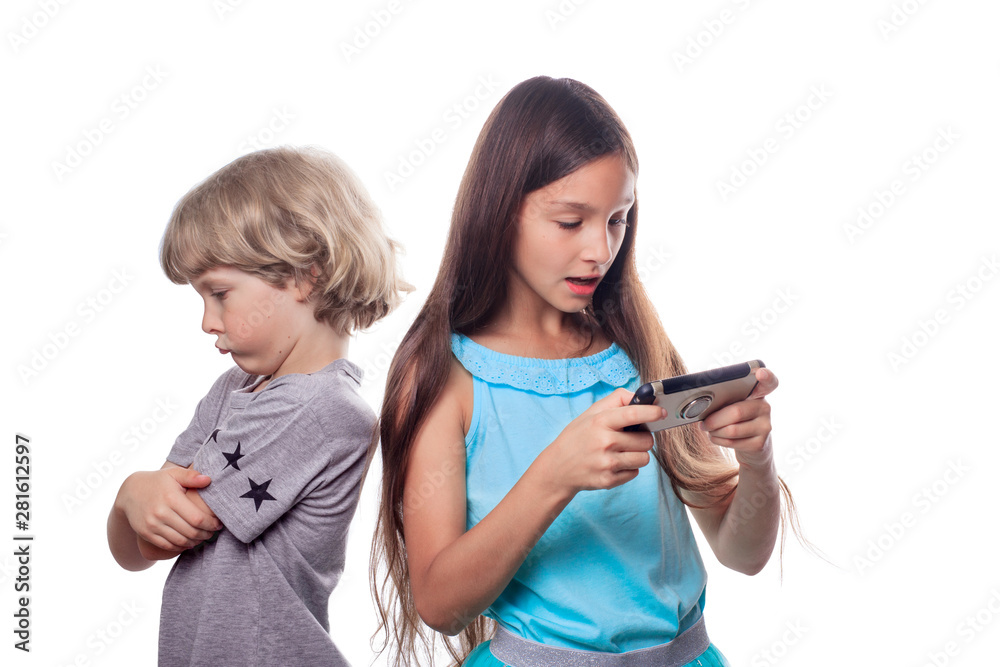 A 10 years old girl with dark hair looking at a mobile phone and a 7 years old blonde boy standing back with an expressive offended face. Siblings resentment and grievance concept A 10 years old girl with dark hair looking at a mobile phone and a 7 years old blonde boy standing back with an expressive offended face. Siblings resentment and grievance concept