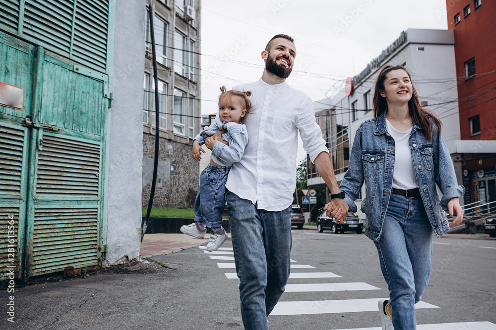 Obraz premium concept of a young happy family with one child: a stylish bearded father, a beautiful young wife and their smiling 2-year-old daughter walking in the city on their hands in the city