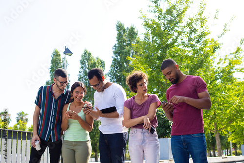 Excited cheerful friends using phones outside. Men and women standing together and showing phone screens to each other. Watching media content concept
