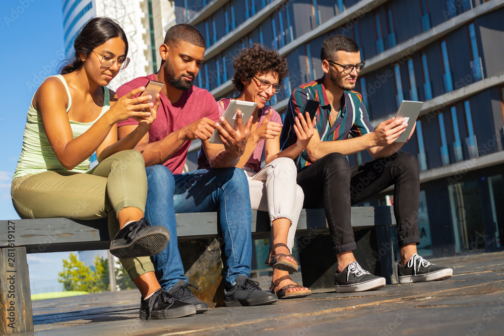 Obraz premium Group portrait of young people focused on their digital gadgets. Men and women sitting on bench outside, using mobile app or consulting internet. Communication concept