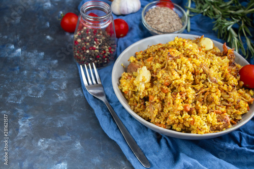 A large plate filled with Uzbek pilaf, next to cherry tomatoes, a dark cloth, fresh pepper in a manually knitted stand. In a transparent dish, spices - zeros and a mixture of peppers. 