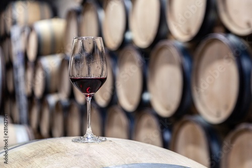 Closeup glass with red wine on background wooden wine oak barrel stacked in straight rows in order, old cellar of winery, vault. Concept professional degustation, winelover, sommelier travel photo