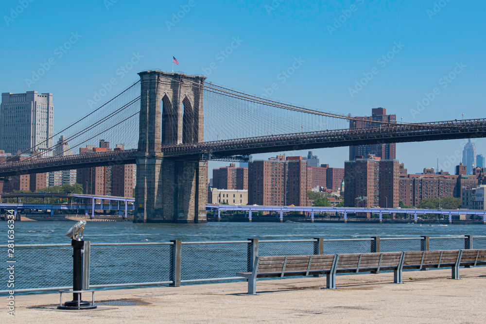 Fototapeta premium Sunny day view of Brooklyn Bridge, American flag, East River and Manhattan Skyline with blue sky