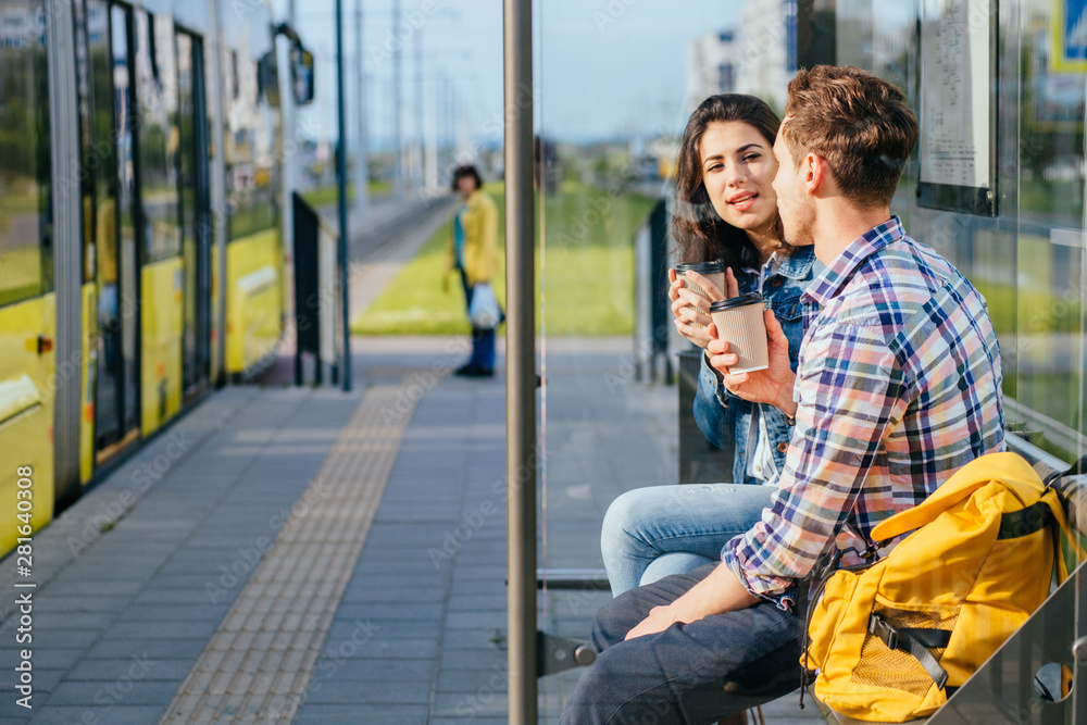 Positive relax couple traveler drinking coffee outdoor at stop while waiting tram, sits on bench at sunny morning.
