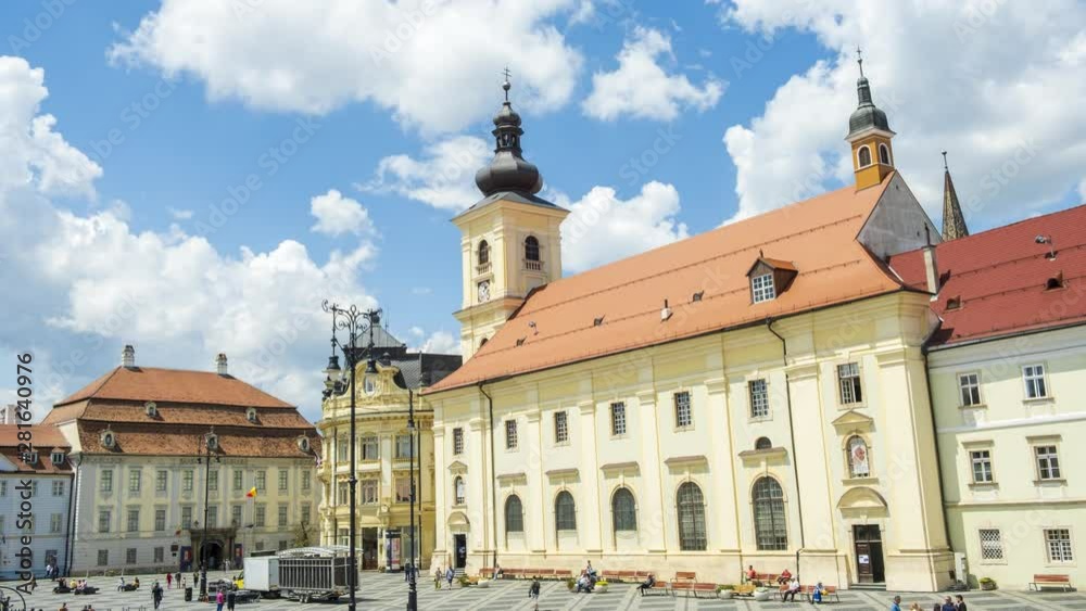Time lapse of Sibiu Main Square during day time, with white clouds passing by fast and their shadows sweeping across the rooftops. Holy Trinity Roman Catholic Church towers visible on the center-right Time lapse of Sibiu Main Square during day time, with white clouds passing by fast and their shadows sweeping across the rooftops. Holy Trinity Roman Catholic Church towers visible on the center-right