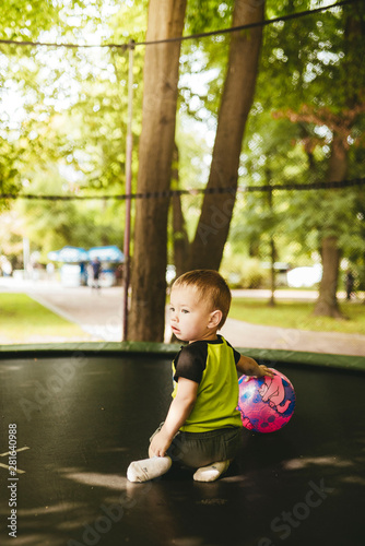 happy and smiling two-year-old boy playing with two balls on a trampoline in the park in summer while walking with his family
