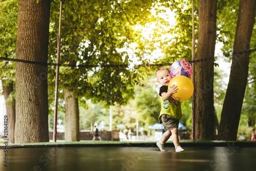 happy and smiling two-year-old boy jumping on a trampoline in the park in summer while walking with his family. green trees and the sun shining through the leaves