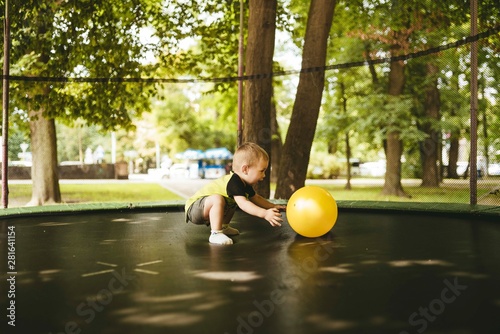 happy and smiling two-year-old boy jumping on a trampoline in the park in summer while walking with his family. green trees and the sun shining through the leaves