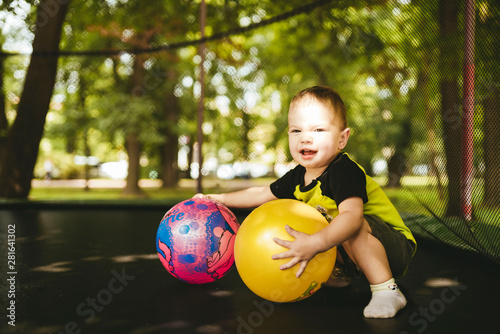 happy and smiling two-year-old boy playing with two balls on a trampoline in the park in summer while walking with his family