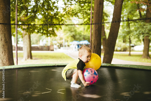 happy and smiling two-year-old boy playing with two balls on a trampoline in the park in summer while walking with his family