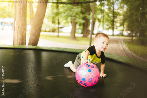 happy and smiling two-year-old boy jumping on a trampoline in the park in summer while walking with his family. green trees and the sun shining through the leaves