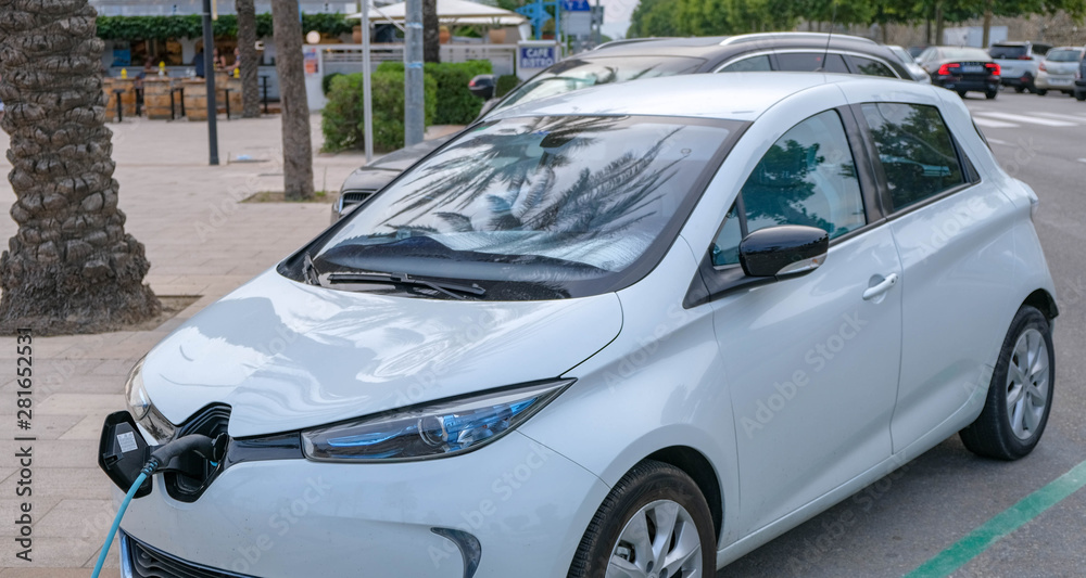 View on white Electric car with the power cable supply plugged. Eco-friendly car on the background of palm trees in summer. Power supply connect to electric vehicle. Side view. Chip electric car. View on white Electric car with the power cable supply plugged. Eco-friendly car on the background of palm trees in summer. Power supply connect to electric vehicle. Side view. Chip electric car.