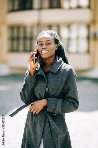 Portrait of young african american girl talking by mobile phone, smiling. Sunny day. Outdoor photo.