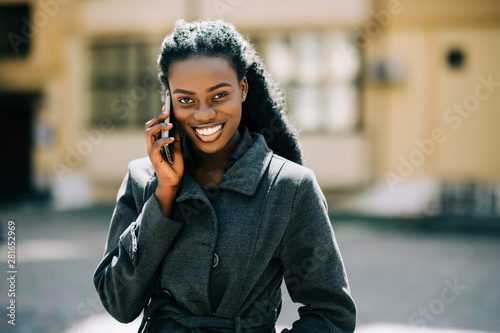 Portrait of young african american girl talking by mobile phone, smiling. Sunny day. Outdoor photo.