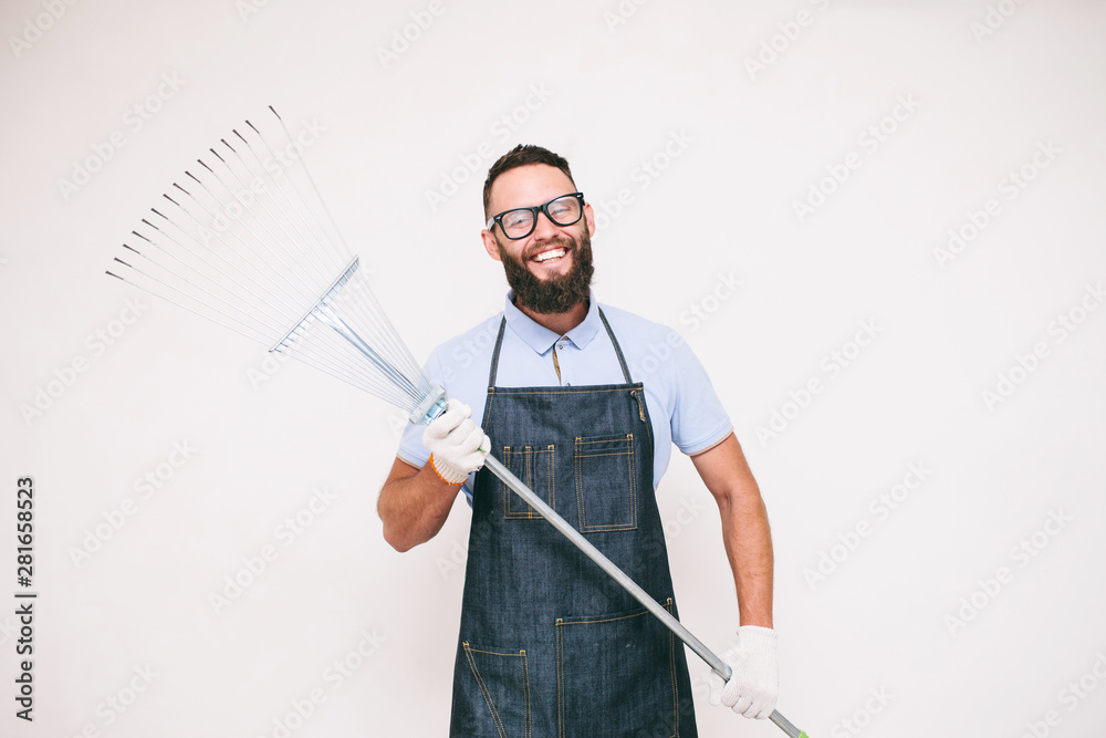 Young joyful emotional bearded gardener in denim apron with instruments. Studio shot on white background