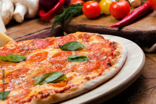 pepperoni pizza decorated with basil on a wooden board, the background is wooden decorated with vegetables and cutlery