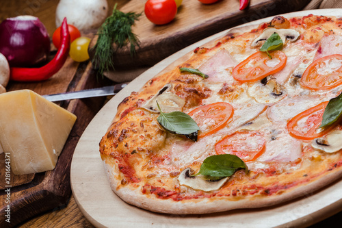 pizza with mushrooms, tomatoes, ham decorated with basil on a wooden board, the background is wooden decorated with vegetables and cutlery