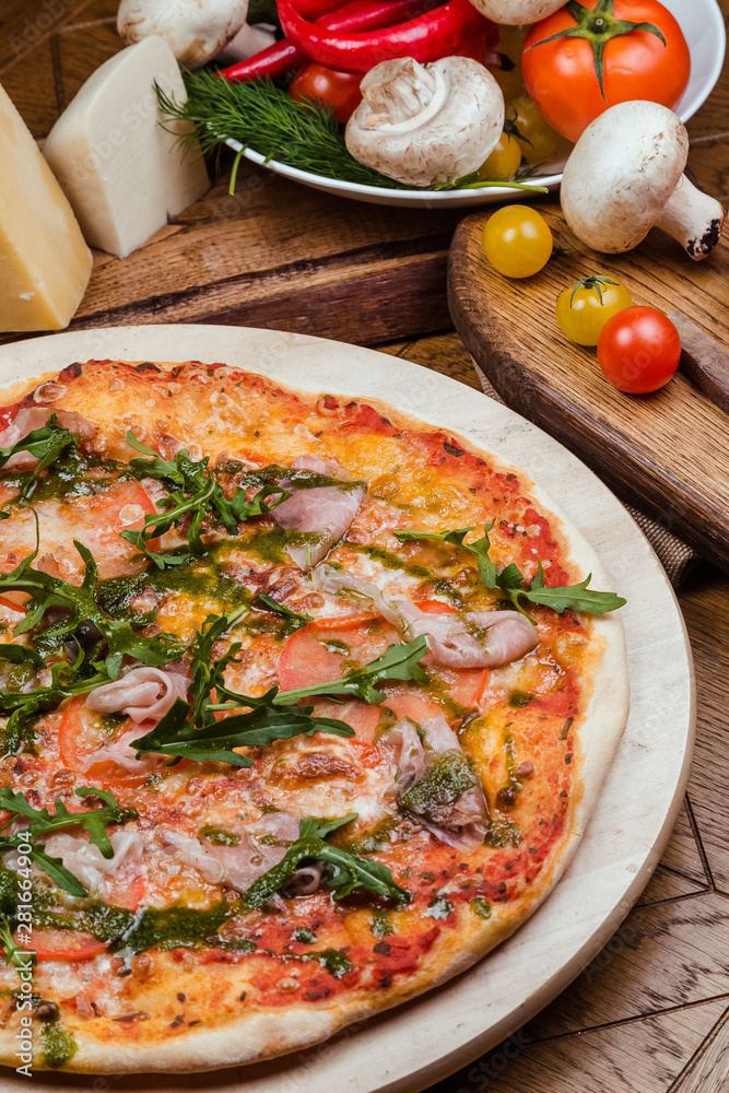 Italian pizza with ham, tomatoes, cheese and arugula on a wooden board, the background is wooden decorated with vegetables and cutlery