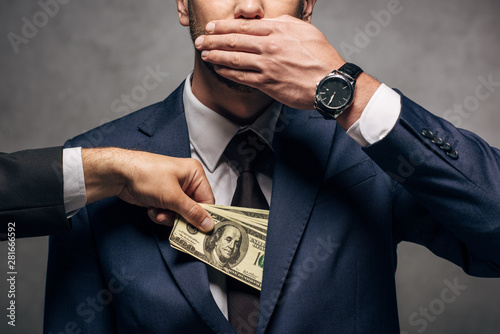 cropped view of man putting cash in pocket of businessman covering face on grey photo