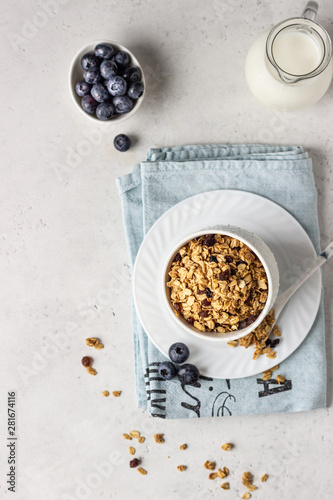 Homemade granola in a white ceramic bowl with raisins, fresh blueberries and milk. Healthy breakfast ingredients on light grey background.