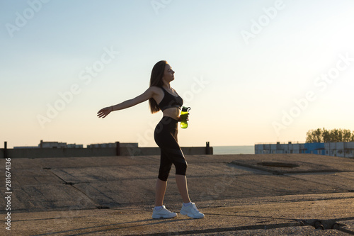Slim athletic girl takes a break between classes and drinks water from a bottle, urban background. Beautiful sky at sunset.  © Mountains Hunter