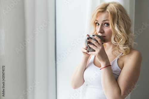 Attractive blonde woman stands near the window with a cup of coffee or tea