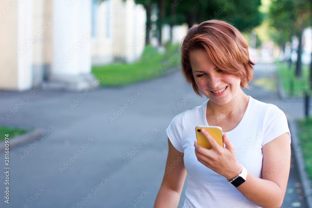 A young happy woman is walking around the city and looking at her phone and smiling. Girl in a white T-shirt and modern technology: smartphone and smartwatch. A young happy woman is walking around the city and looking at her phone and smiling. Girl in a white T-shirt and modern technology: smartphone and smartwatch.