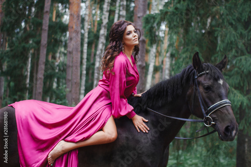 Portrait of young beautiful smiling brunette woman wearing pink silk dress riding dark horse at summer green forest.
