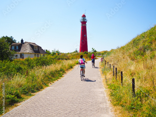 Canvas-taulu Back view cyclists traveling on the road heading towards to lighthouse in Schiermonnikoog islands, on sand dune against blue sky
