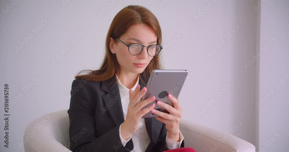 Closeup portrait of young cheerful businesswoman in glasses typing on the tablet looking ta camera smiling happily sitting in the armchair Closeup portrait of young cheerful businesswoman in glasses typing on the tablet looking ta camera smiling happily sitting in the armchair