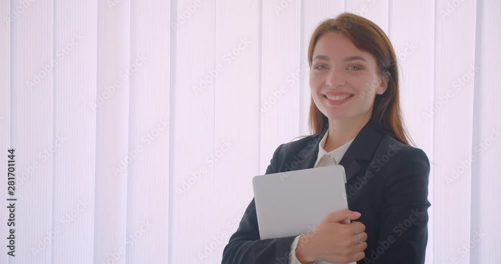 Closeup portrait of young caucasian businesswoman holding the laptop looking at camera smiling happily standing indoors in the white office Closeup portrait of young caucasian businesswoman holding the laptop looking at camera smiling happily standing indoors in the white office