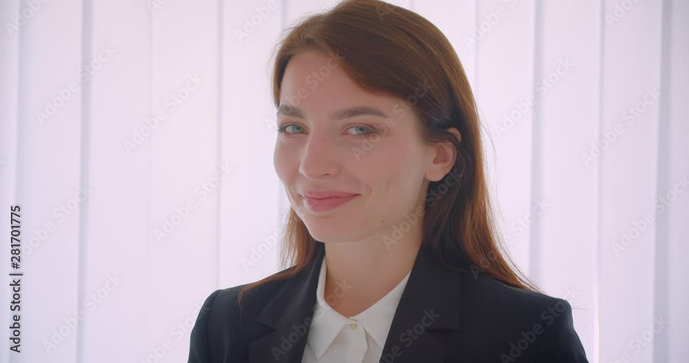 Closeup portrait of young successful businesswoman looking at camera smiling happily standing indoors in the white office Closeup portrait of young successful businesswoman looking at camera smiling happily standing indoors in the white office