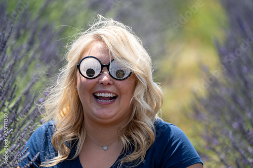 Crazy Blonde woman wearing googly eyes novelty sunglasses while sitting in a field of lavender, looking surprised with her mouth open