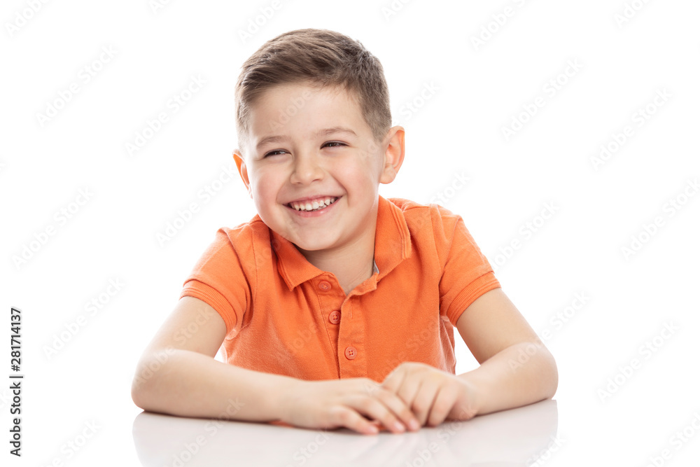 Laughing school-age boy in a bright orange polo t-shirt sits at a table. Close-up. Isolirvoan on a white background. Laughing school-age boy in a bright orange polo t-shirt sits at a table. Close-up. Isolirvoan on a white background.
