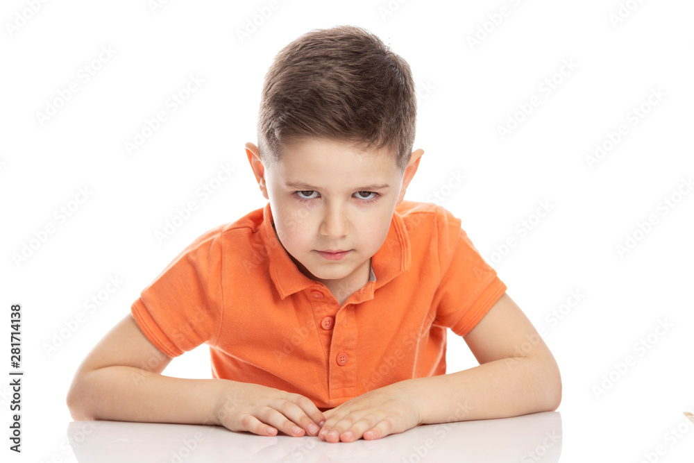 Frowning serious school-age boy in a bright orange polo t-shirt sits at a table. Close-up. Isolirvoan on a white background. Frowning serious school-age boy in a bright orange polo t-shirt sits at a table. Close-up. Isolirvoan on a white background.