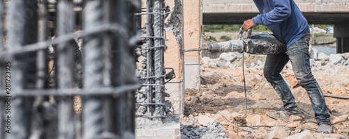 Construction worker holding and using pneumatic jackhammer drill equipment to break reinforced concrete pillar at road worl construction site.