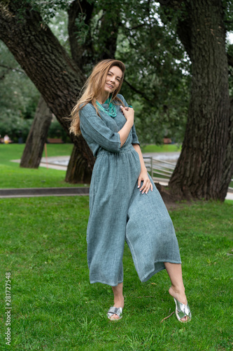 young white girl, natural appearance, long hair, beautiful face, green linen light jumpsuit. Cool pose in full growth in city park near trees. comfortable linen womens clothing.