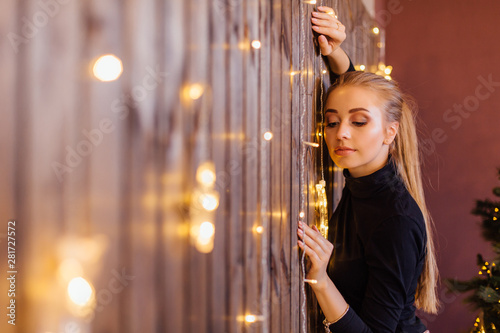 Beautiful young woman dressed in a sweater and skirt standing next to the wooden wall with star and moon lights