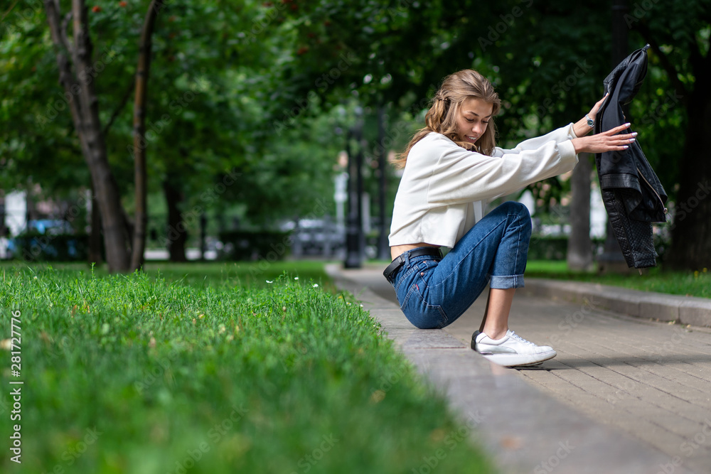 Fototapeta premium beautiful slender European girl sat down on curb in city park. Caucasian girl sitting on curb sidewalk. Lifestyle, fashionable look. Great mood.