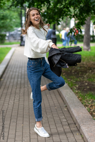 beautiful young European girl is having fun walking along pavement in city park. Happy smile caucasian girl. Lifestyle, pullover, jeans, sneakers. Waving arms, wide step, go fast.