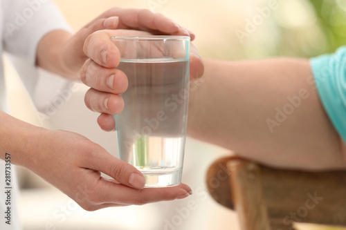 Nurse giving glass of water to elderly man indoors, closeup. Assisting senior generation