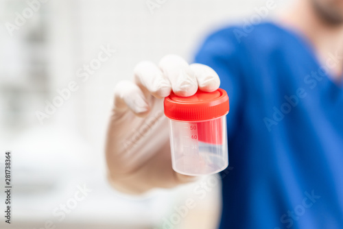 doctor in blue uniform and latex gloves is holding an empty plastic container for taking urine samples, light background. Medical concept.
