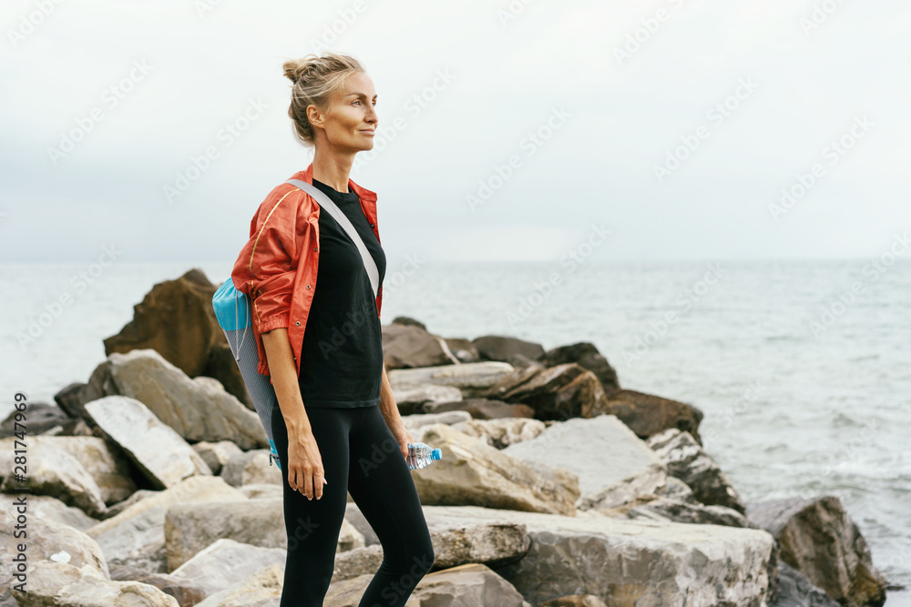 An athletic Caucasian woman of 30 after jogging stands on a breakwater from large cobblestones and looks into the distance, holds a water bottle and a backpack with a sports mat on her back. An athletic Caucasian woman of 30 after jogging stands on a breakwater from large cobblestones and looks into the distance, holds a water bottle and a backpack with a sports mat on her back.