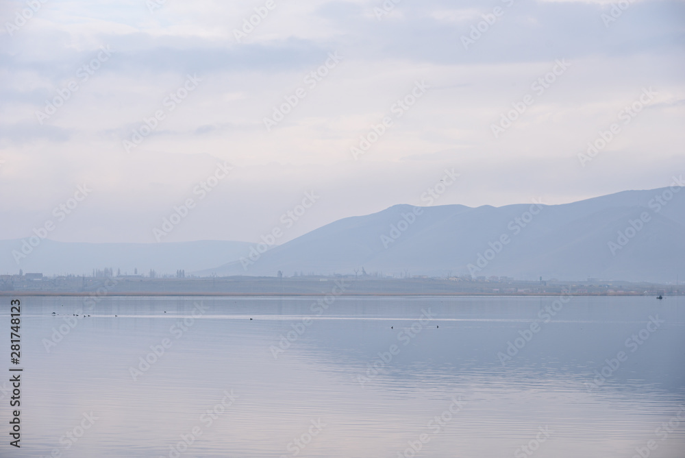 Water expanses of the endless Lake Sevan in the foreground with grass and two fishermen and ducks on a cloudy day with mountain views in the background.