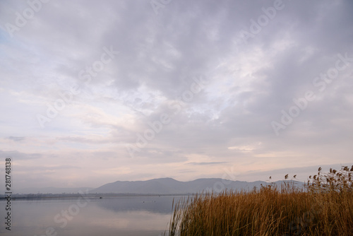 Water expanses of the endless Lake Sevan in the foreground with grass and two fishermen and ducks on a cloudy day with mountain views in the background.