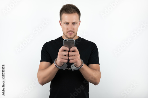 handsome young man in black t-shirt holding smartphone while being handcuffed on isolated white background © studioprodakshn